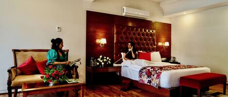 A hotel room with a wooden floor, a sofa with red cushions, a bed with a floral bedspread, and a nightstand with a lamp.