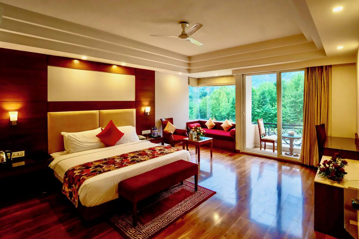 A spacious bedroom with a large bed, a sitting area with a sofa, and a balcony with a view of trees.