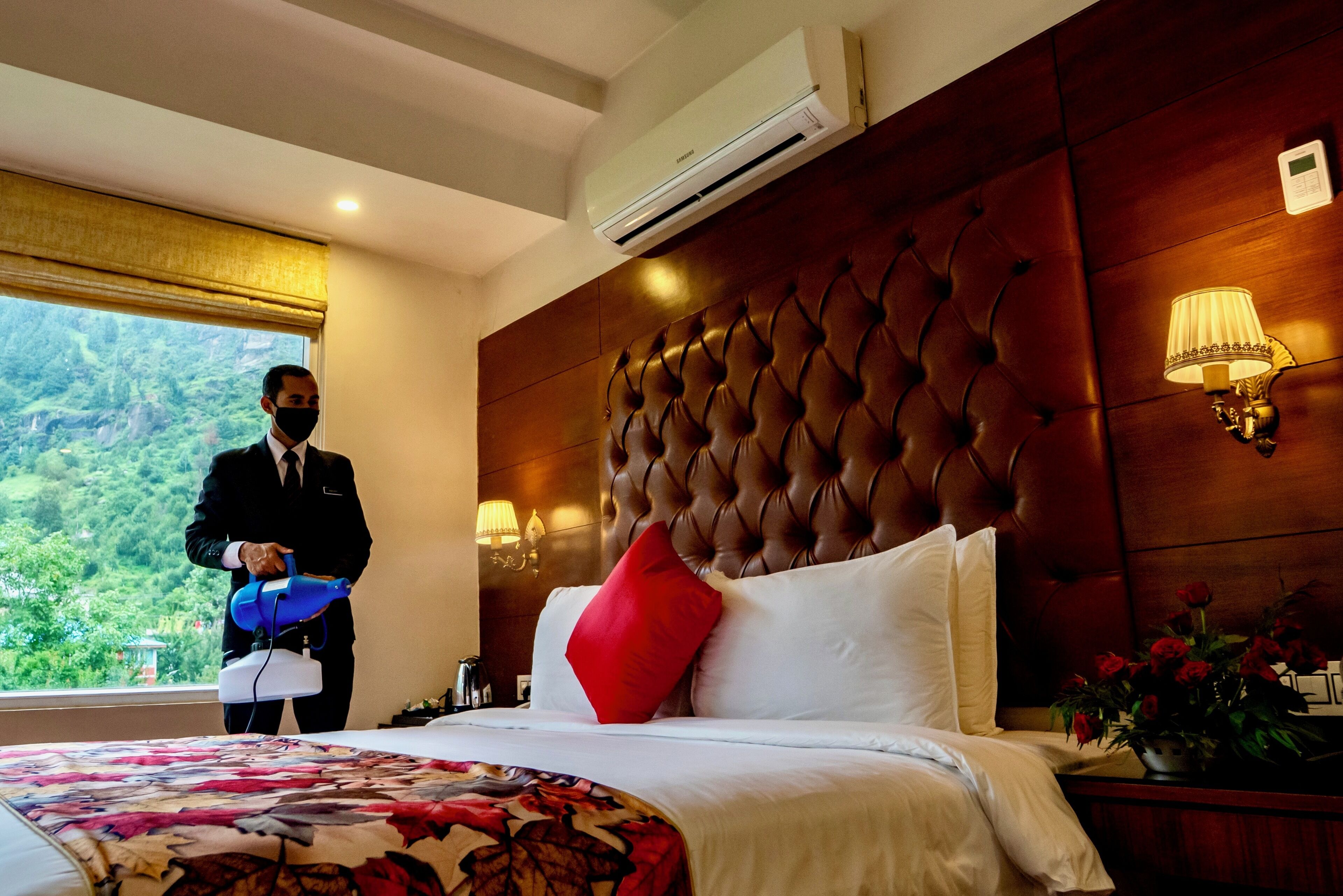 Une chambre d’hôtel avec un grand lit, un oreiller rouge, une couvre-lit à motifs floraux, une tête de lit en bois et une fenêtre donnant sur un paysage verdoyant.