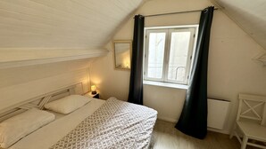 Quarto pequeno e acolhedor com teto inclinado, cama com roupa de cama estampada, mesa de cabeceira com abajur, janela com cortinas e uma cadeira branca.