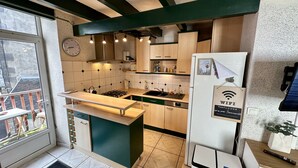 Cozinha compacta com ilha central, armários de madeira e fogão embutido. Um relógio está fixado na parede, e um sinal de WiFi aparece na geladeira.