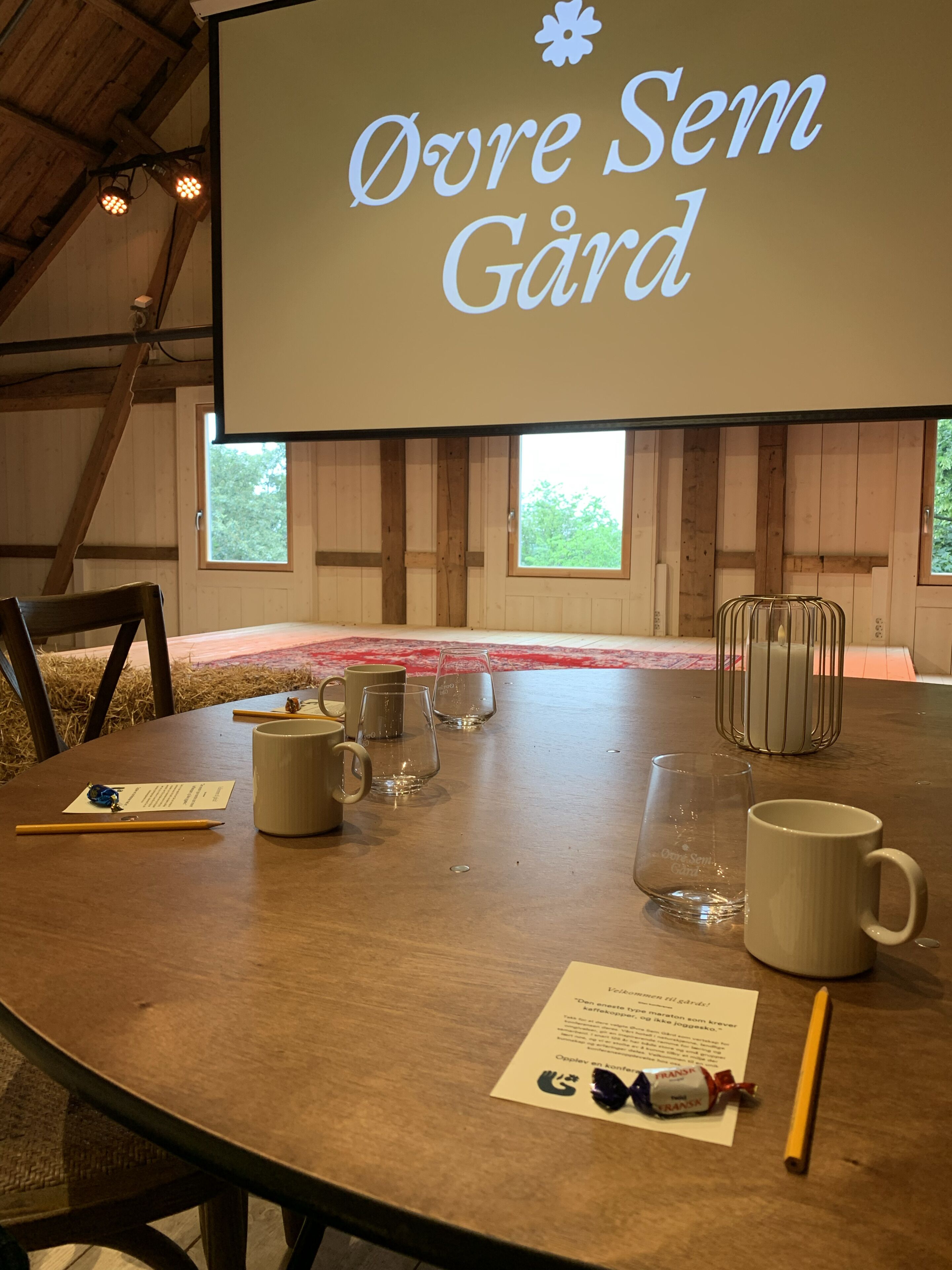 Un salón de conferencias con una pantalla grande que muestra 'Øvre Sem Gard', una mesa de madera con sillas y varios objetos como tazas y vasos.