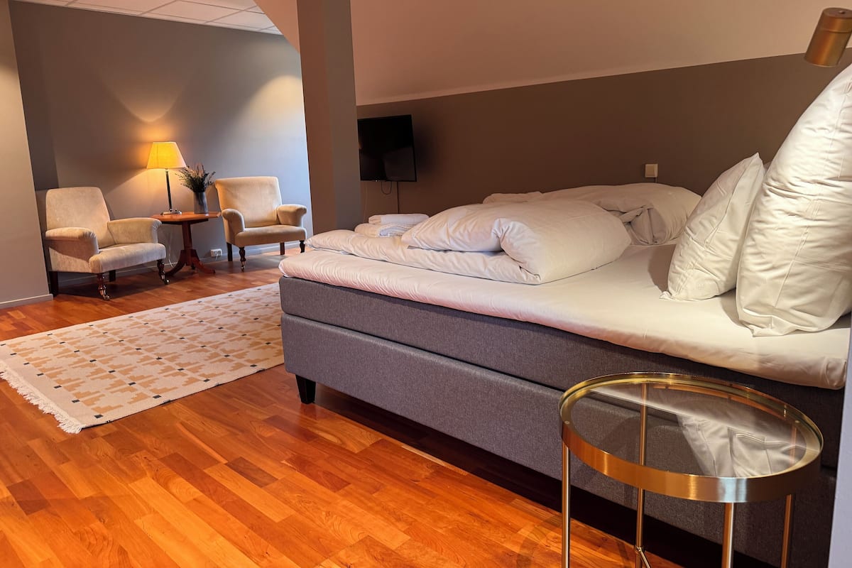 Una habitación de hotel moderna con una cama grande, dos sillones, una mesita y un televisor.