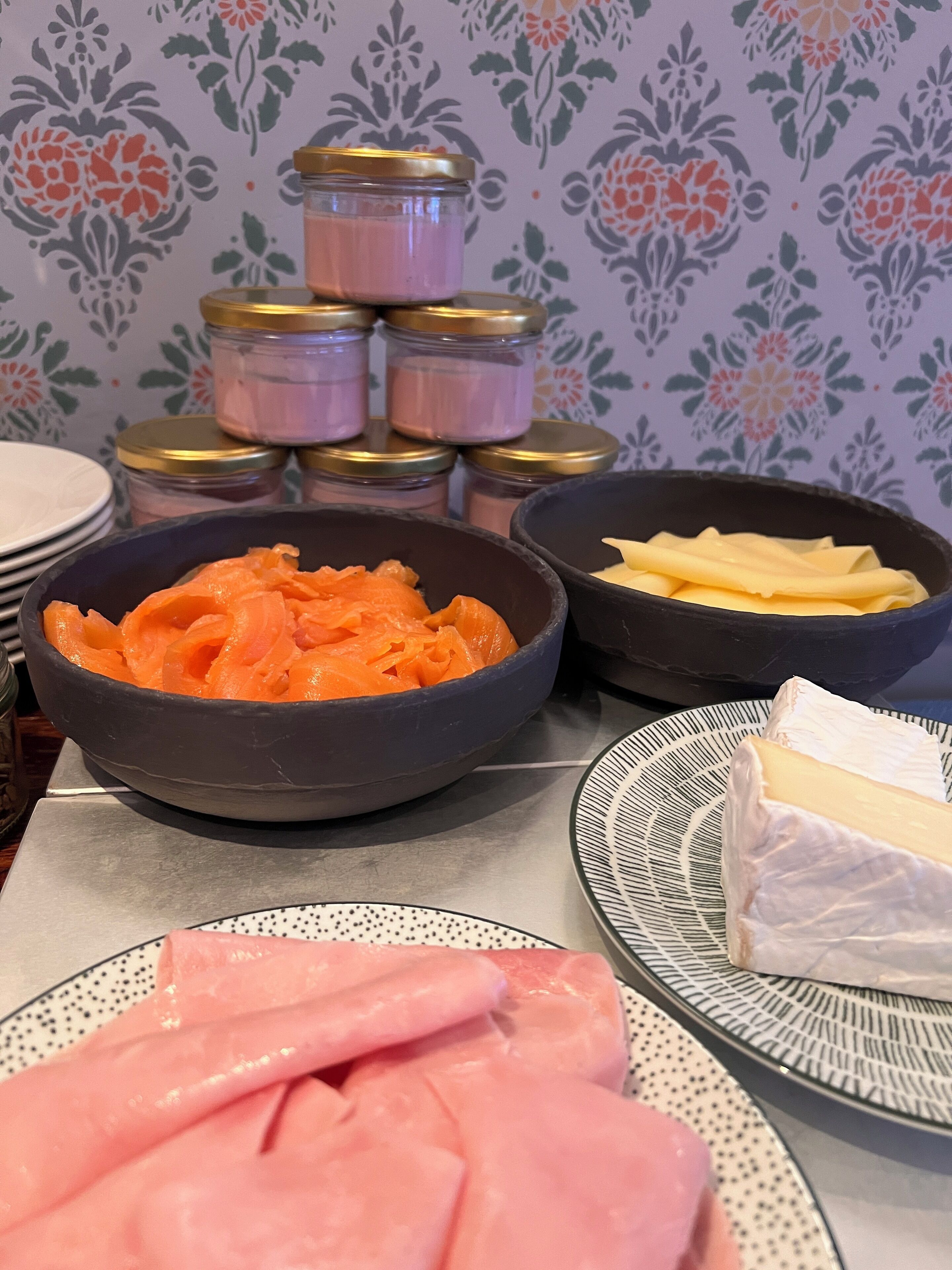 A table with various food items including bowls of sliced meat, cheese, and a stack of jars with lids.
