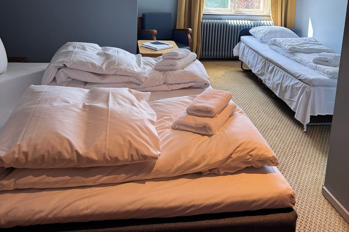 Una habitación de hotel con dos camas, una ventana con cortinas, un radiador y una pequeña mesa con un libro.