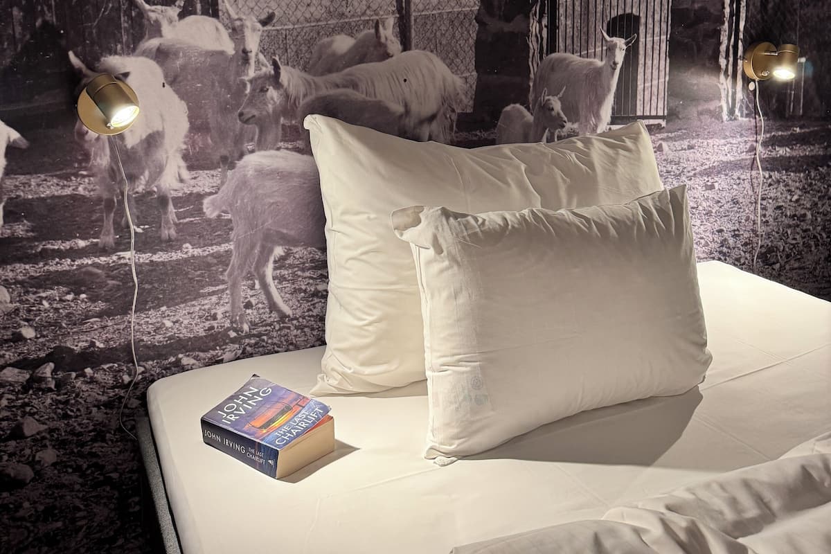 Un dormitorio con cama, almohadas y un libro. Se aprecia un mural en la pared con animales y un granero.