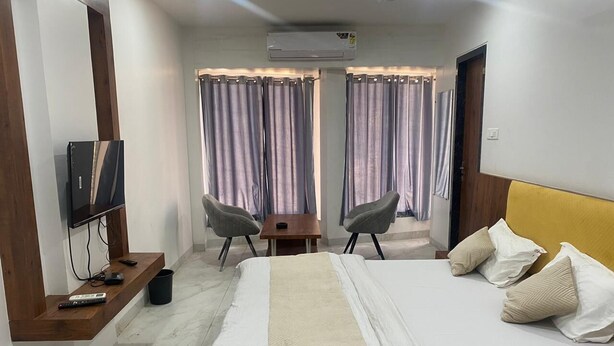 Une chambre d’hôtel avec un lit, un téléviseur fixé sur une étagère en bois, un bureau avec des chaises et des rideaux.