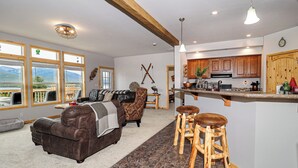 A spacious living room with a brown leather couch, a patterned armchair, and a wooden bar stool.