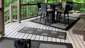 Una terraza de madera con zona de estar cubierta, una parrilla y un perro acostado sobre una alfombra.