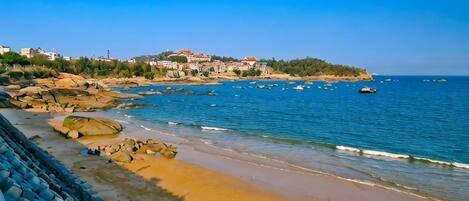 Escena de playa costera con costa rocosa, cielo azul despejado y una vista distante de un pueblo con edificios.