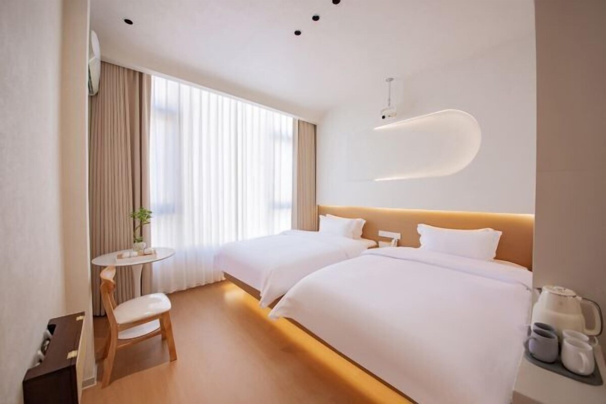 Ein Hotelzimmer mit zwei Betten, einem kleinen runden Tisch mit einem Stuhl, einer Wandleuchte und einem Fenster mit Vorhängen.