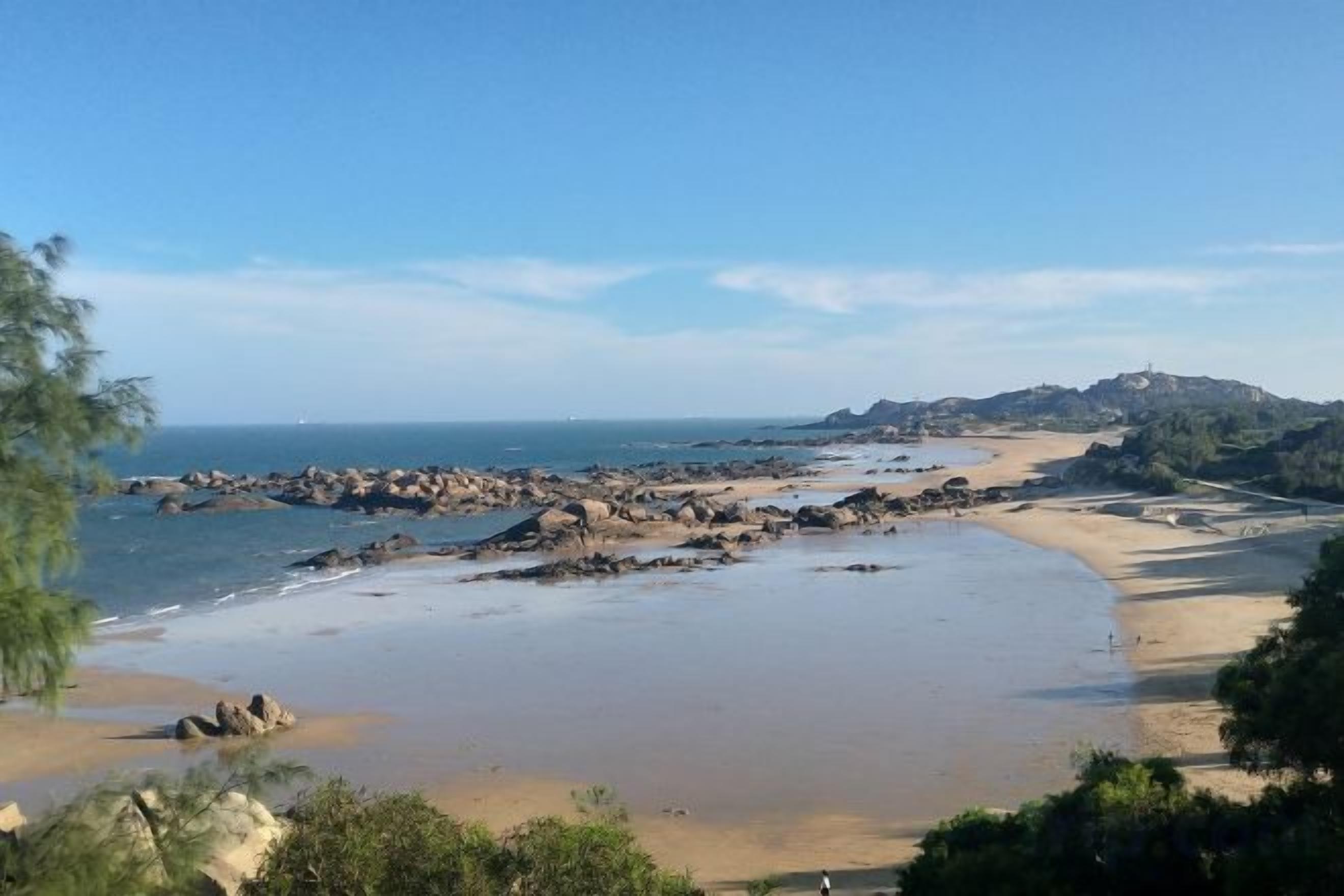 Un paisaje costero con una playa arenosa, afloramientos rocosos y una colina distante.