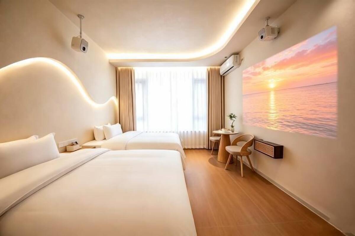 Hotellihuone, jossa on kaksi sänkyä, työpöytä, tuoli, suuri ikkuna ja seinällä auringonlaskua kuvaava taulu.