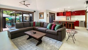 A modern living room with a sectional sofa, a coffee table, and a glass table. There is a kitchen area with red cabinets and a dining area with a bar. - Banyak Villa (Horseshoe Bay)