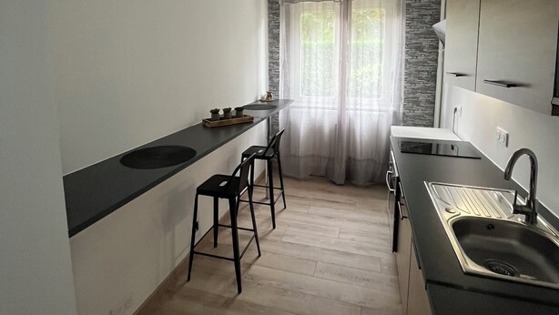 Eine moderne Küche mit schwarzem Arbeitsplatz, Spüle aus Edelstahl und Holzboden. Zwei Barhocker und ein Fenster mit leichtem Vorhang sind ebenfalls vorhanden.