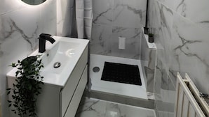 Ein modernes Badezimmer mit Marmorboden, einem runden Spiegel, einer Dusche mit Glastür und einem weißen Waschbecken mit schwarzem Wasserhahn.