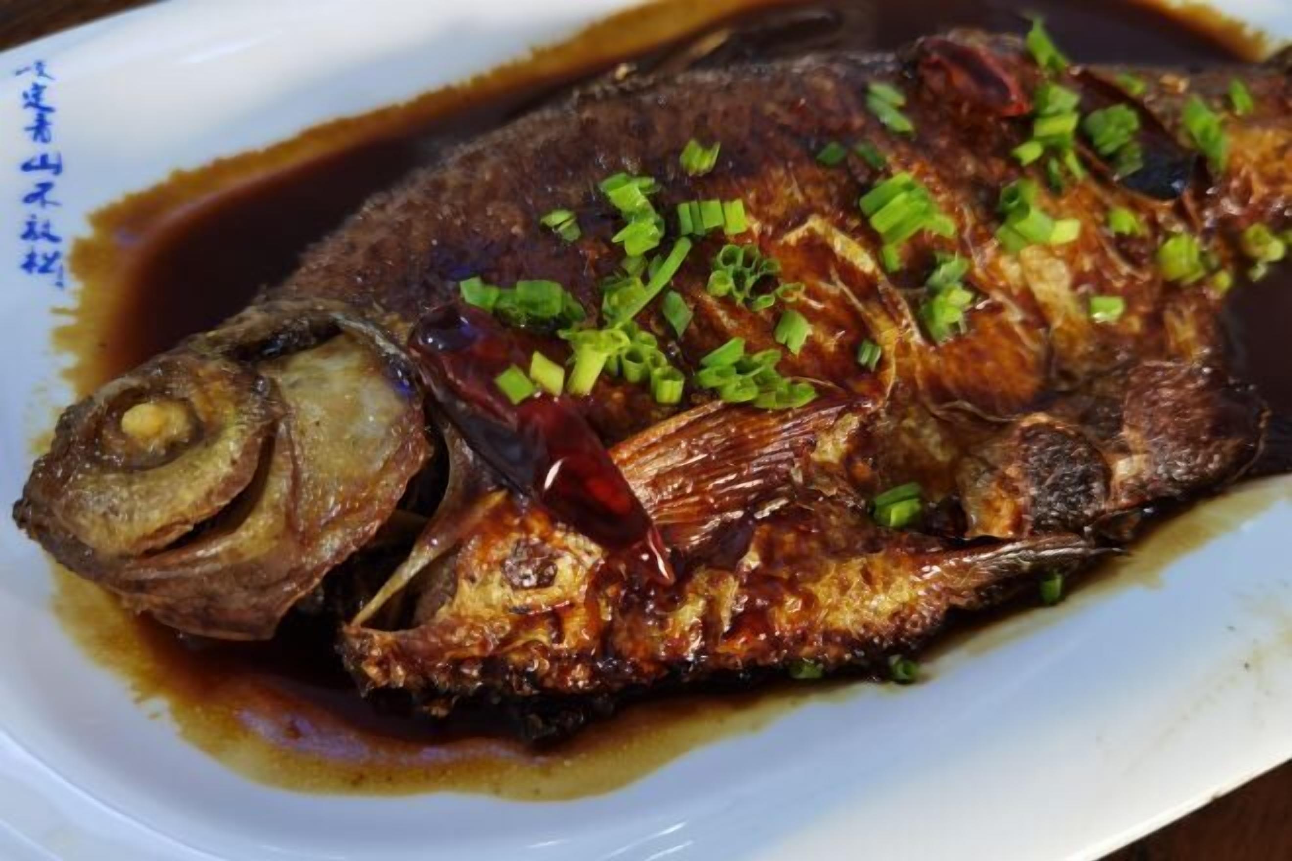 A whole fried fish with a crispy skin, garnished with chopped green onions, served on a white plate with a dark sauce.