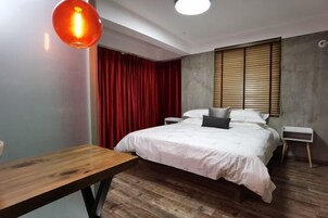Un dormitorio con suelo de madera, una cama con ropa de cama blanca, una cortina roja, un cabecero de madera y una mesita de noche pequeña.