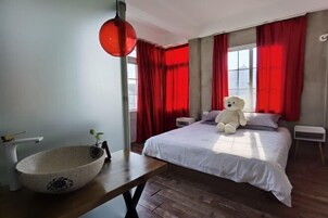 Un dormitorio con una cama, una mesita de noche, una cortina roja, un peluche de oso blanco y un cuenco sobre una mesa de madera.