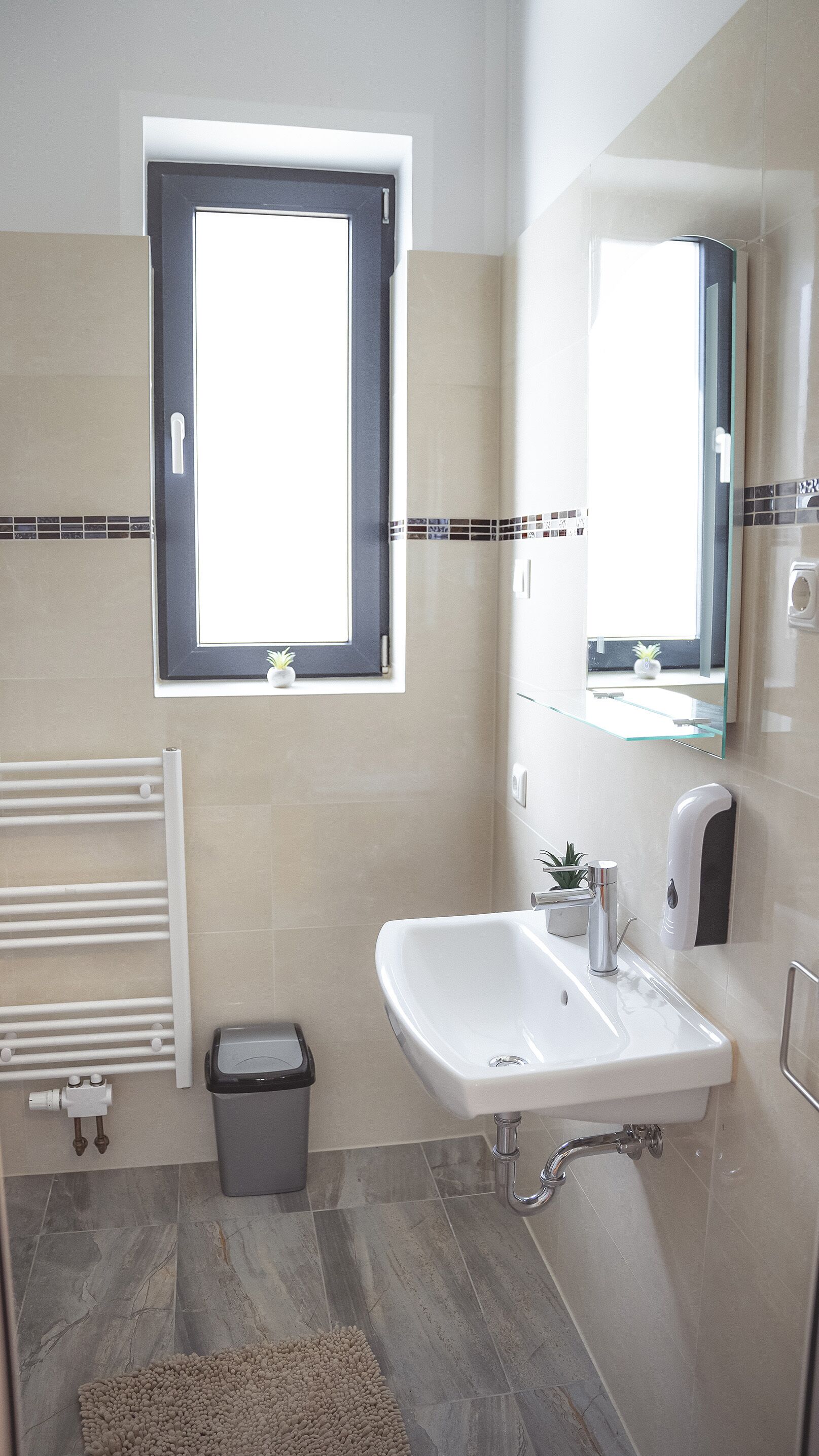 A modern bathroom with a large window, a white sink, a mirror, a towel rack, and a small trash bin.