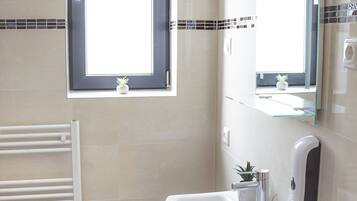 A modern bathroom with a large window, a white sink, a mirror, a towel rack, and a small trash bin.