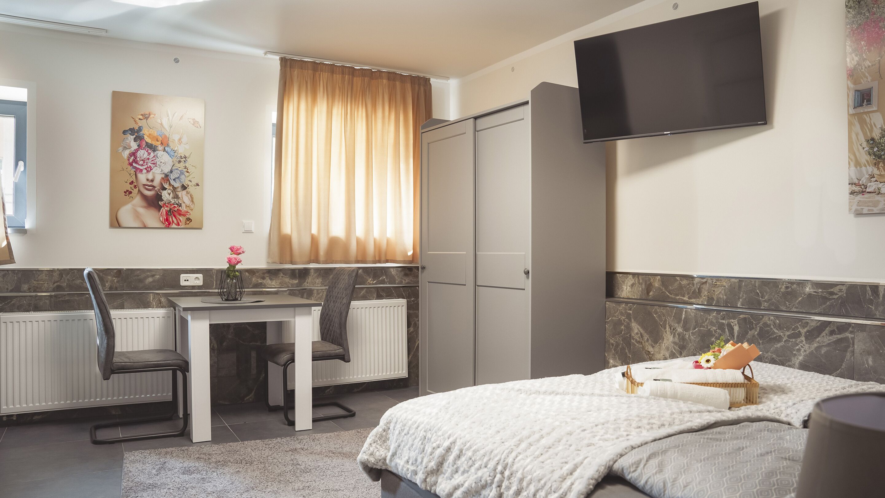 Habitación de hotel con cama, escritorio con silla, radiador, televisor montado en la pared y ventana con cortinas.
