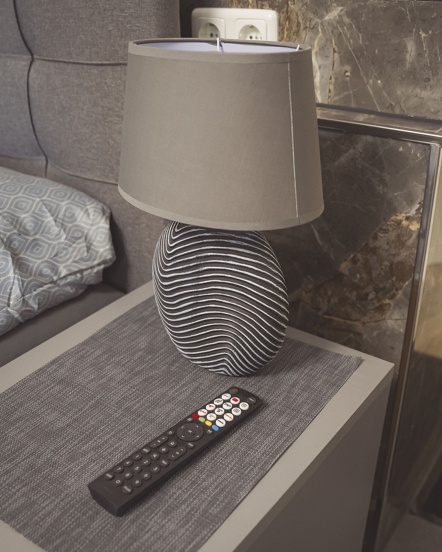 A modern lamp with a unique design on a bedside table, a remote control, and a patterned pillow on a couch.