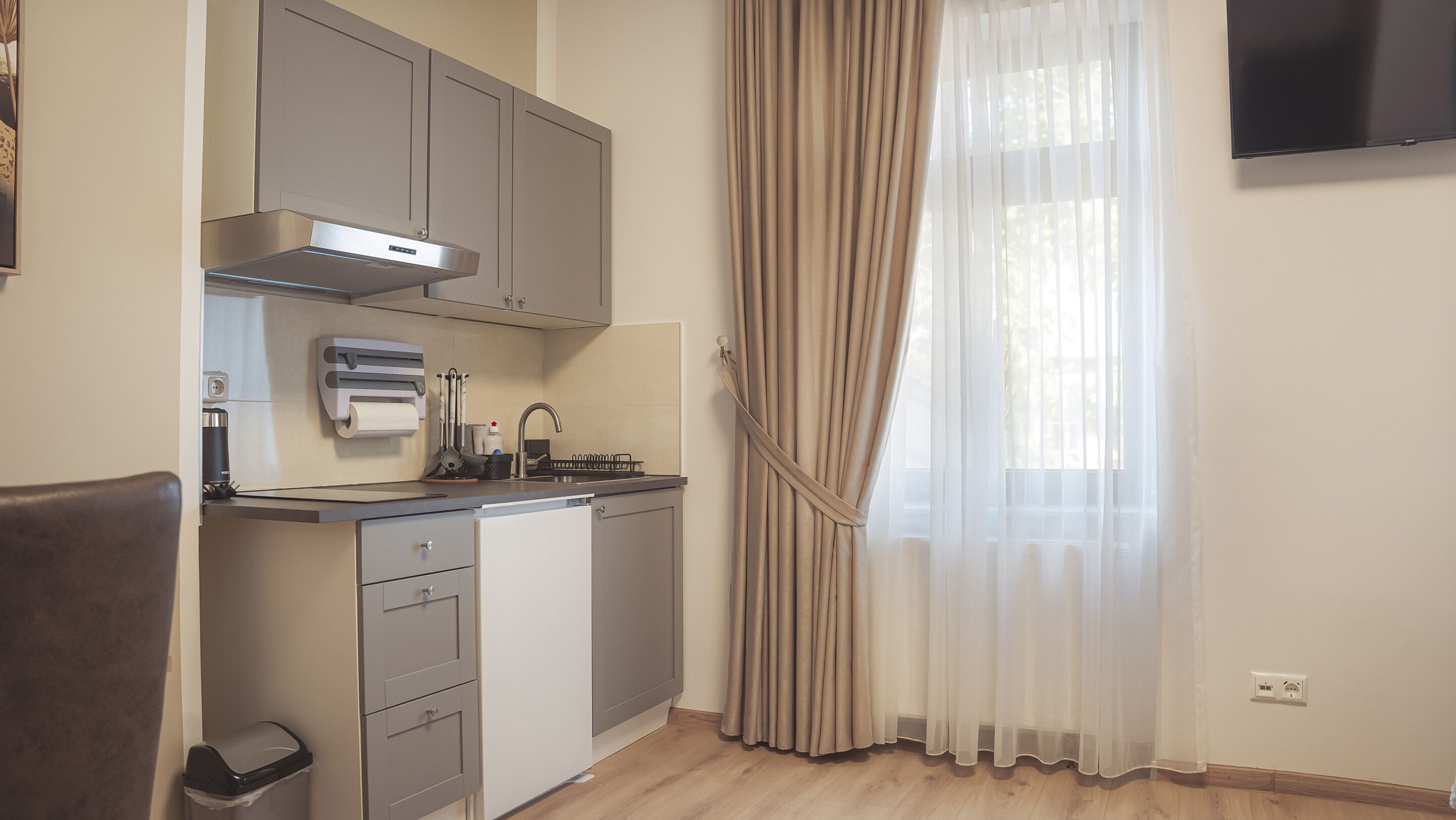 Cuina compacta amb rentaplats, fogó i rentamans. Hi ha una finestra amb cortines translúcides i un televisor de pantalla plana penjat a la paret.