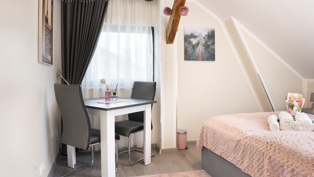 A bedroom with a bed, a desk, and a chair. There is a window with curtains, a picture on the wall, and a trash can.