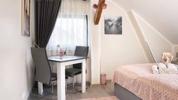 A bedroom with a bed, a desk, and a chair. There is a window with curtains, a picture on the wall, and a trash can.