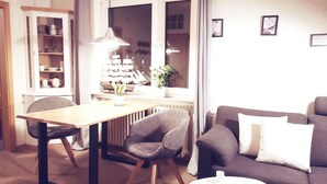 A cozy living room with a dining table, a sofa, and a chair. There is a window with curtains, a radiator, and a plant on the windowsill. - Apartment in Esens, Deutschland (Esens)