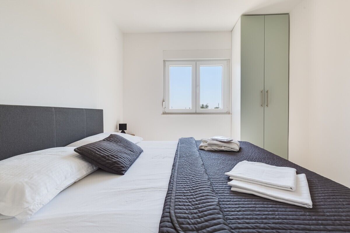 A neatly made bed with a grey headboard, a window above it, and a green wardrobe.