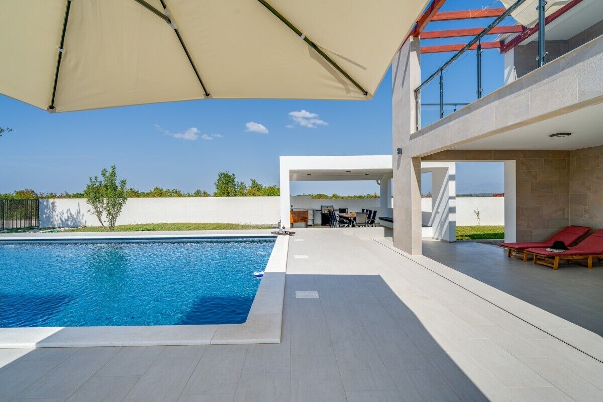 Un área de piscina exterior moderna con una piscina azul cristalina, un espacio de estar cubierto y vistas al paisaje circundante.