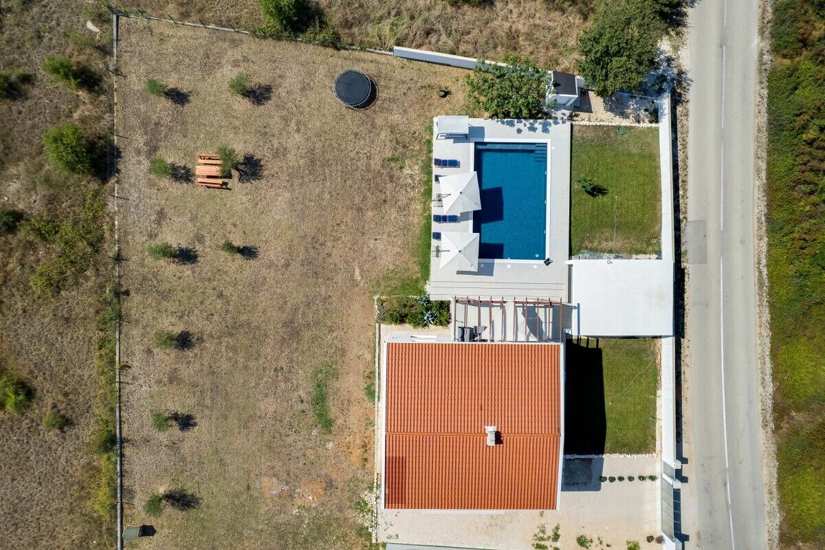 Eine Vogelperspektive zeigt ein Anwesen mit Schwimmbad, überdachter Terrasse und großzügiger, offener Fläche mit vereinzelten Bäumen.