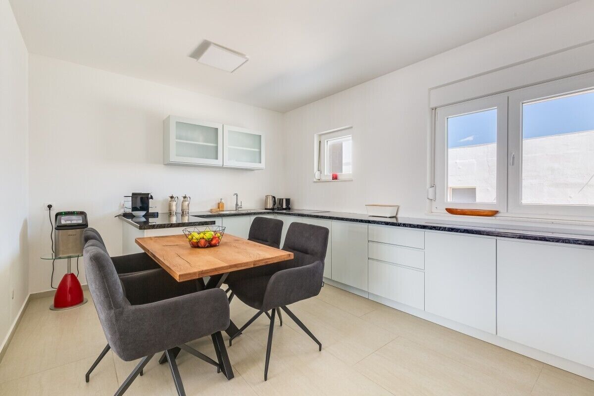 A modern kitchen with white cabinets, a wooden dining table with four chairs, a window with a view, and a small window above the sink.