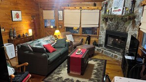 Un interior de cabaña acogedor con suelo de madera, chimenea, sofá, sillón mecedora y mesa de comedor.