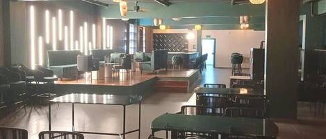 A spacious restaurant with high tables and chairs, a stage for performances, and ambient lighting.