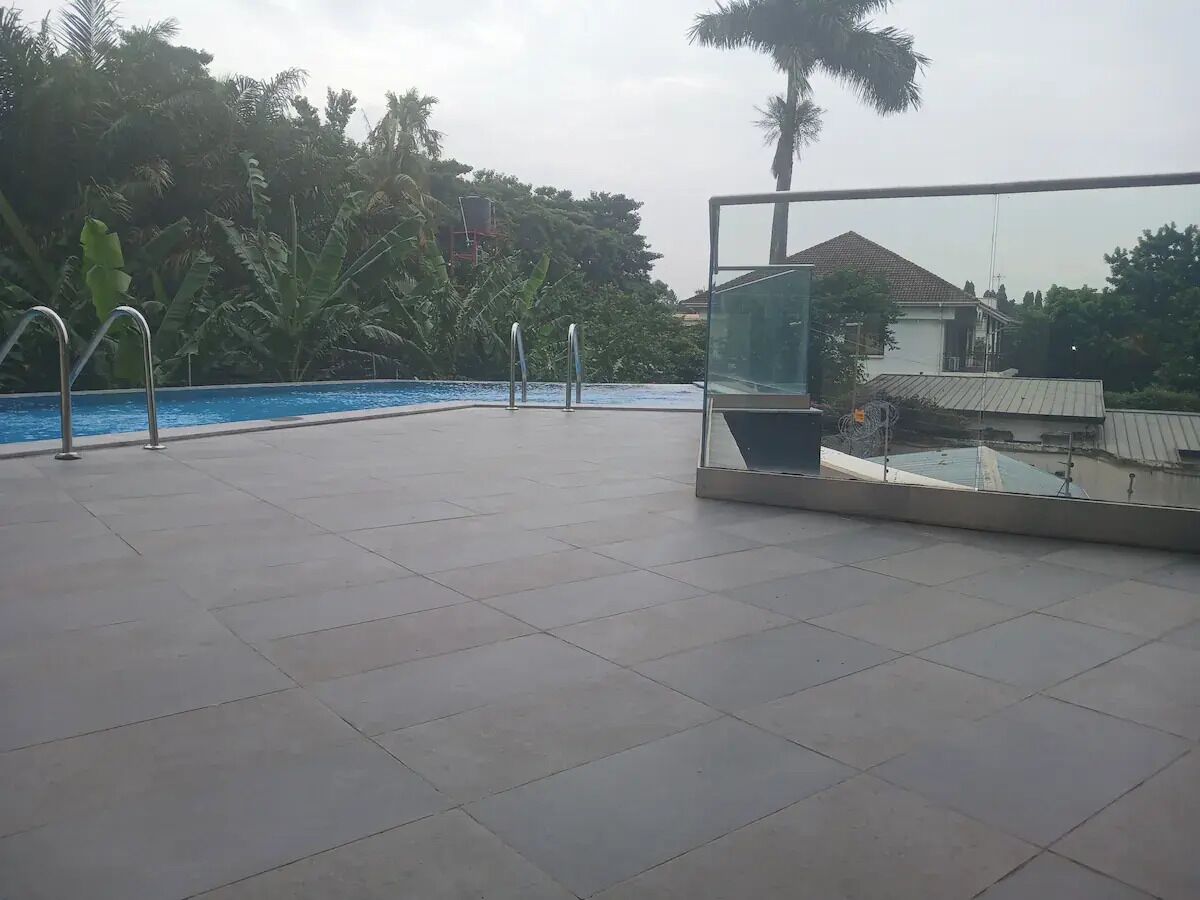 Een zwembad op het dak, voorzien van betegelde vloeren, metalen zwemladder en een glazen balustrade met uitzicht op een woongebied met bomen en een huis.