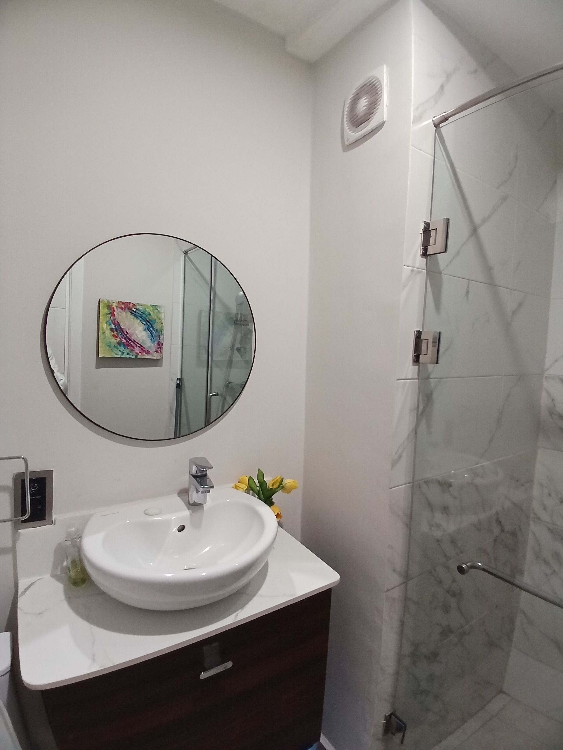 A bathroom with a white sink, a round mirror, a colorful artwork, and a glass shower enclosure.