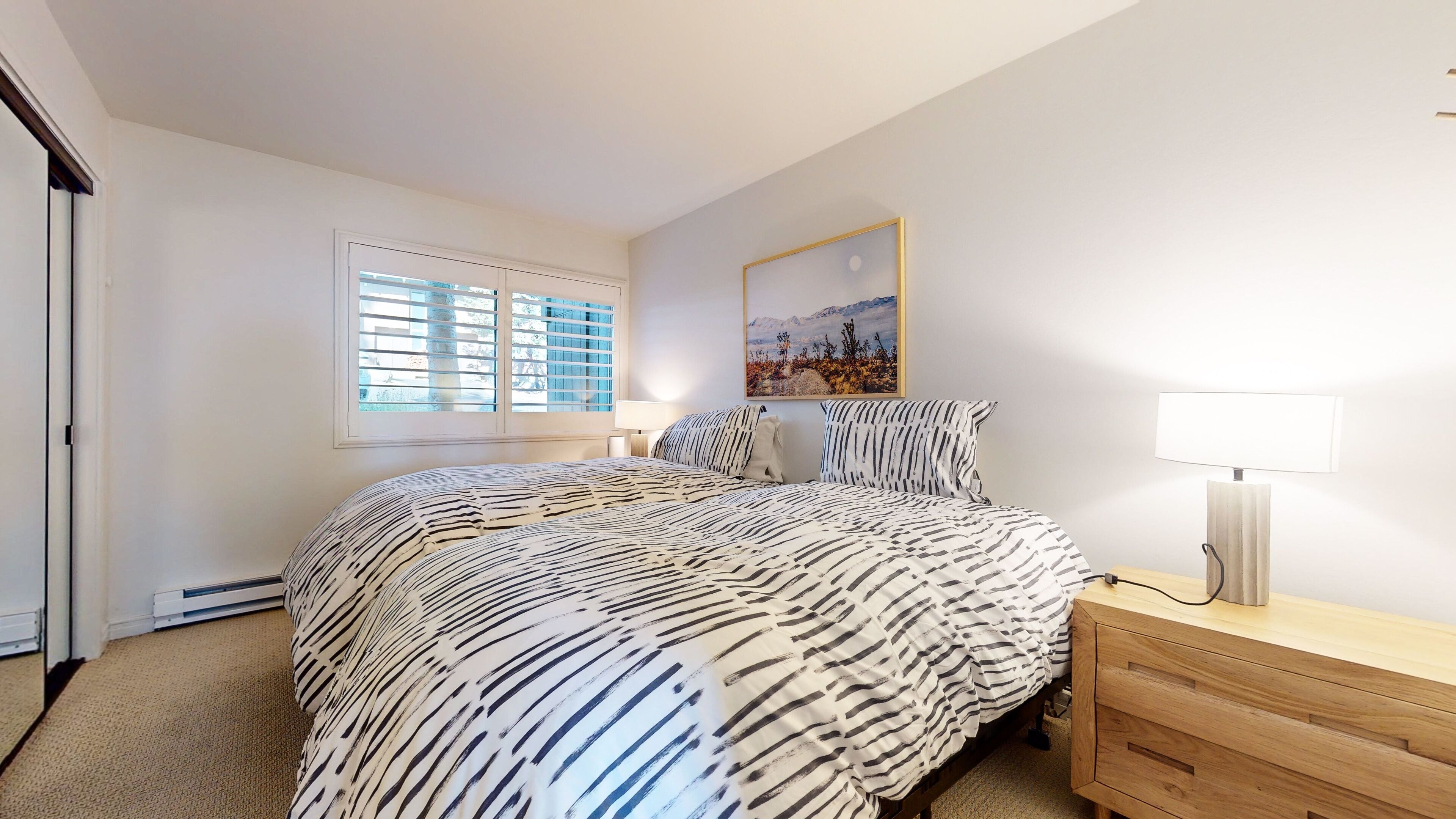 Ein Schlafzimmer mit einem Bett, einem Nachttisch, einer Lampe, einem Fenster mit Jalousien und einem Gemälde an der Wand.