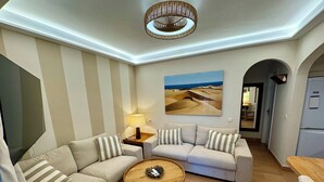 Un salon moderne avec un canapé d’angle, une table basse ronde en bois et une lampe. Un tableau représentant un paysage désertique est accroché au mur.