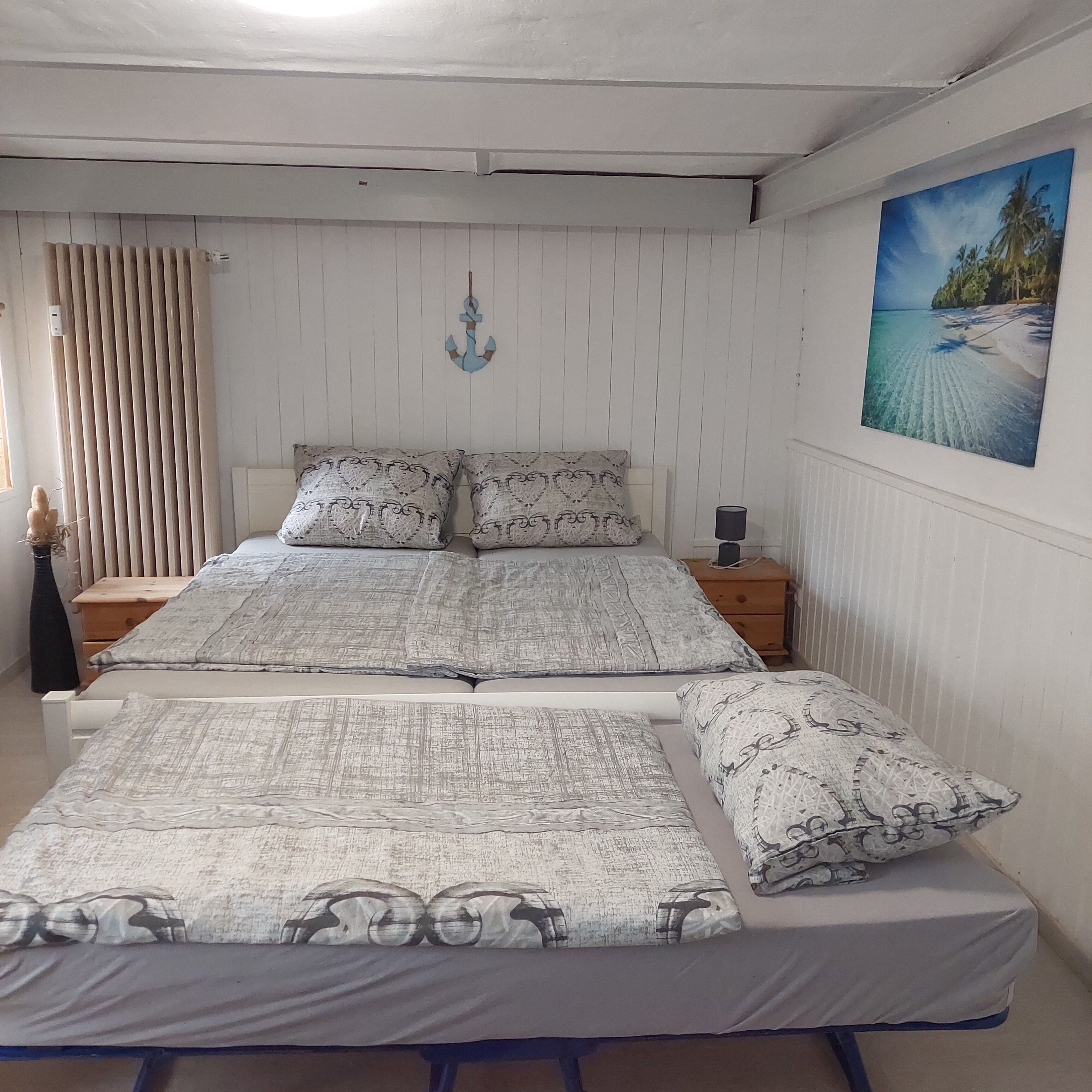 Una cama doble con ropa de cama estampada, una mesita de noche con lámpara, un aparador de madera, una ventana con cortinas y un adorno colgado en la pared.