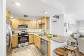 A modern kitchen with wooden cabinets, stainless steel appliances, a granite countertop, and a dining area with a sofa and bar stools. - Penthouse Beachfront Condo | Pool/Fitness/Grills (Marco Island)