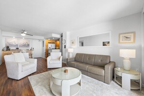 Un salon moderne avec une table basse en verre, un canapé beige et un fauteuil blanc. On aperçoit à l’arrière-plan une cuisine avec des meubles en bois et un coin repas comprenant une table et des chaises.
