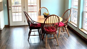 Zona pranzo con tavolo e sedie in legno, un lampadario e vista esterna attraverso ampie finestre.