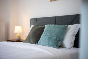 A neatly made bed with white bedding, a grey upholstered headboard, and two pillows, one green and one white. - Alex’s 2-Bedroom Apartment in Athens (Greece - Athens)