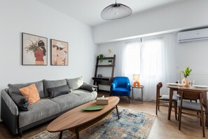 A modern living room with a grey sofa, a wooden coffee table, a blue armchair, and a dining area with a table and chairs. - Alex’s 2-Bedroom Apartment in Athens (Greece - Athens)