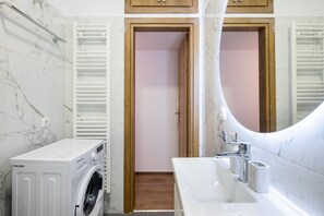 A modern bathroom with a marble wall, a washing machine, a sink with a mirror, and wooden cabinets. - Alex’s 2-Bedroom Apartment in Athens (Greece - Athens)