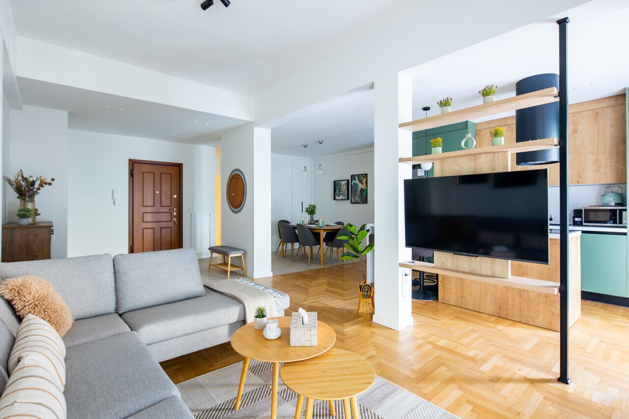 Ein modernes Wohnzimmer mit einem grauen Sofa, einem runden Holztisch und einem Fernseher. Im Hintergrund befindet sich ein Essbereich mit Tisch und Stühlen.