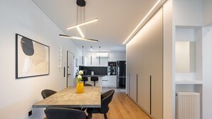 A modern kitchen with white cabinets, a black refrigerator, and a wooden dining table with chairs. - Bloom - Luxury 2-Bedroom Apartment in Athens (Greece - Glyfada)